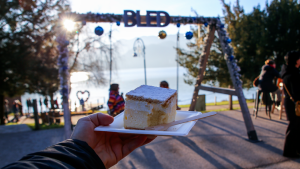 Person holding up a serve of Bled Cream Cake in front of Lake Bled in Slovenia by Hera Sparnon