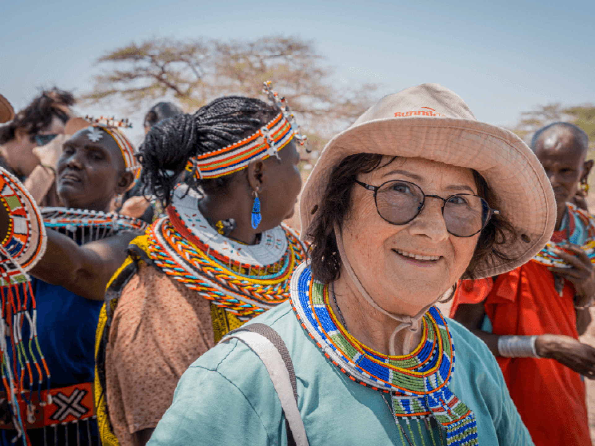 Marion Bunnik wearing a traditional blue and green beaded necklace with a group of women from the Samburu Unity Village in Shaba, Kenya captured by Amie Bunnik. The women are each wearing multi-coloured beaded necklaces and head pieces.